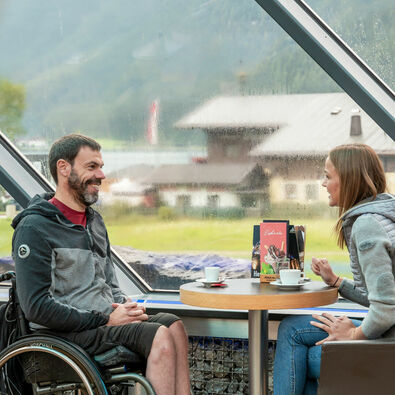 Barrierefreiheit am Achensee Zwei Freunde bei denen einer im Rollstuhl sitzt unterhalten sich im Cafe Vitalberg mit einem heißen Getränk bei Regenwetter.